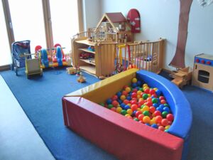 La foto muestra un fragmento del área para niñas y niños con una piscina de bolas, un rincón de juego vallado, dos cochecitos para muñecas, dos balancines de elefante y una casa de muñecas.